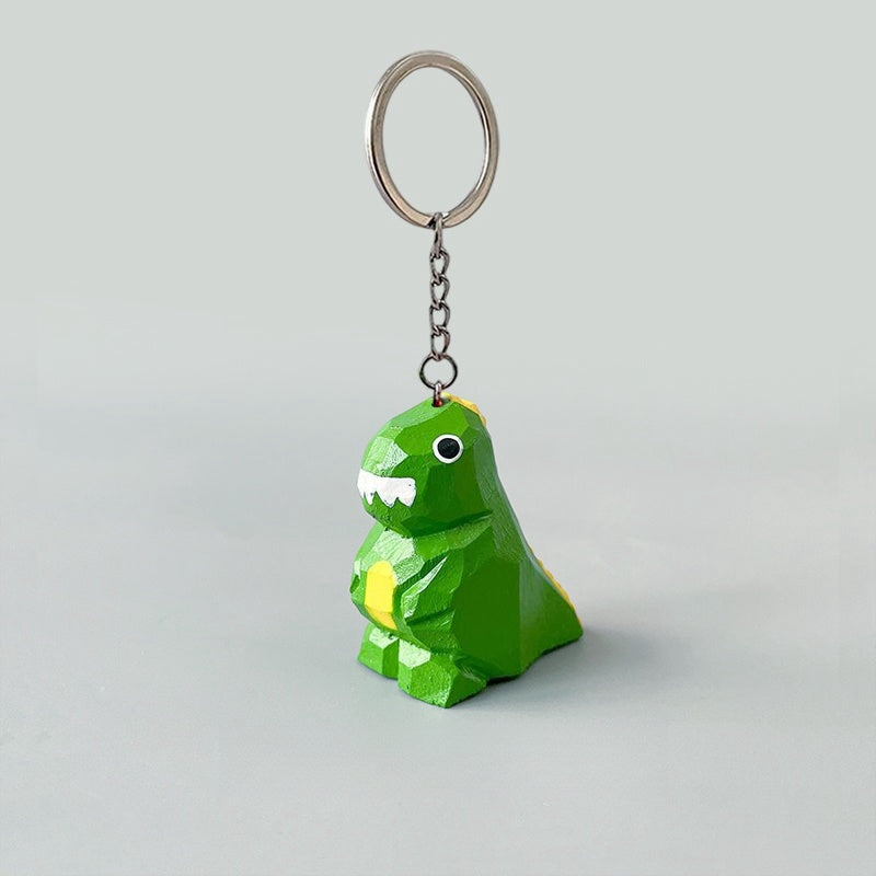 Green dinosaur keychain with white teeth and yellow belly, hanging from a keyring against a light gray background.