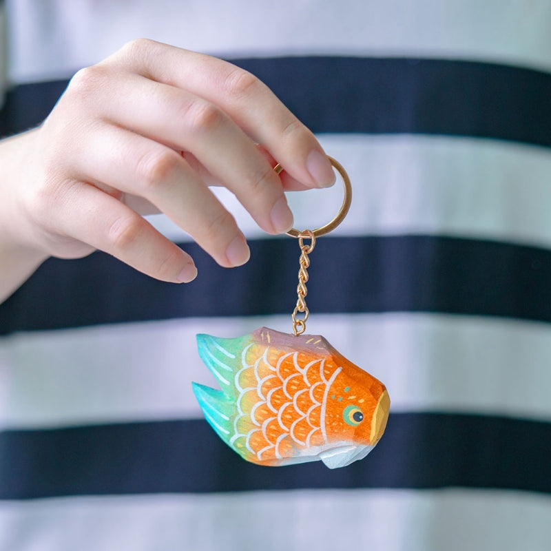 Hand displaying a colorful fish keychain with bright orange and green scales, with a striped shirt in the background.