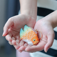 Two hands gently cupping a colorful fish keychain with bright orange and green scales, showcasing its unique and vibrant appearance.