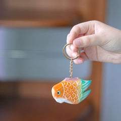 Hand holding a colorful fish keychain with bright orange and green scales, against a blurred background.