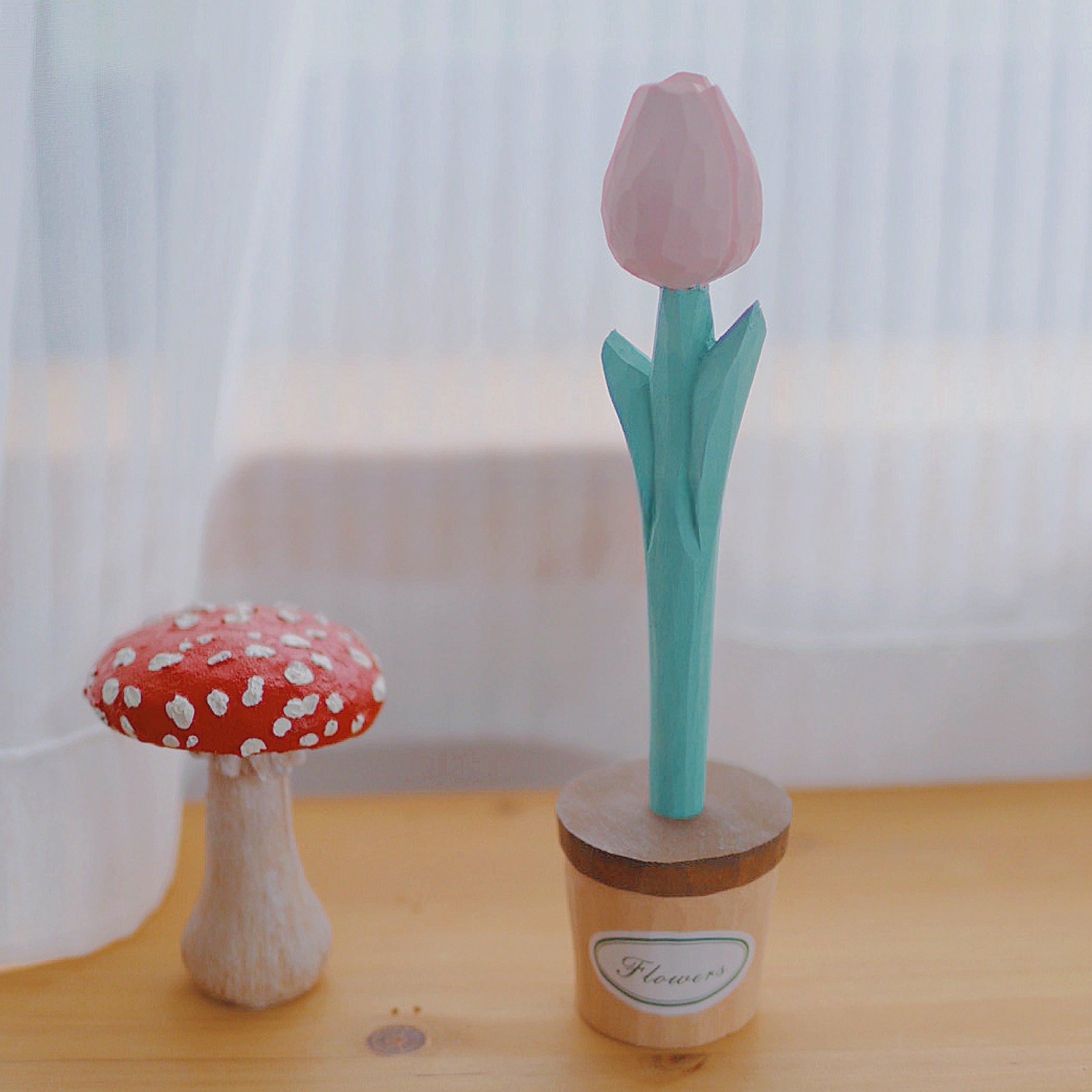 A light pink flower pen in a small pot labeled "Flowers," placed on a wooden table next to a red mushroom decoration.