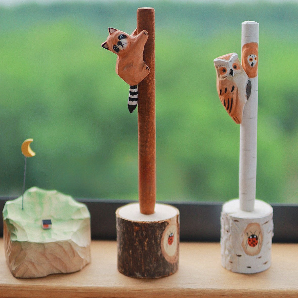 Wooden pens with hand-carved raccoon and owl figures displayed in tree stump holders, with a miniature landscape decoration nearby.