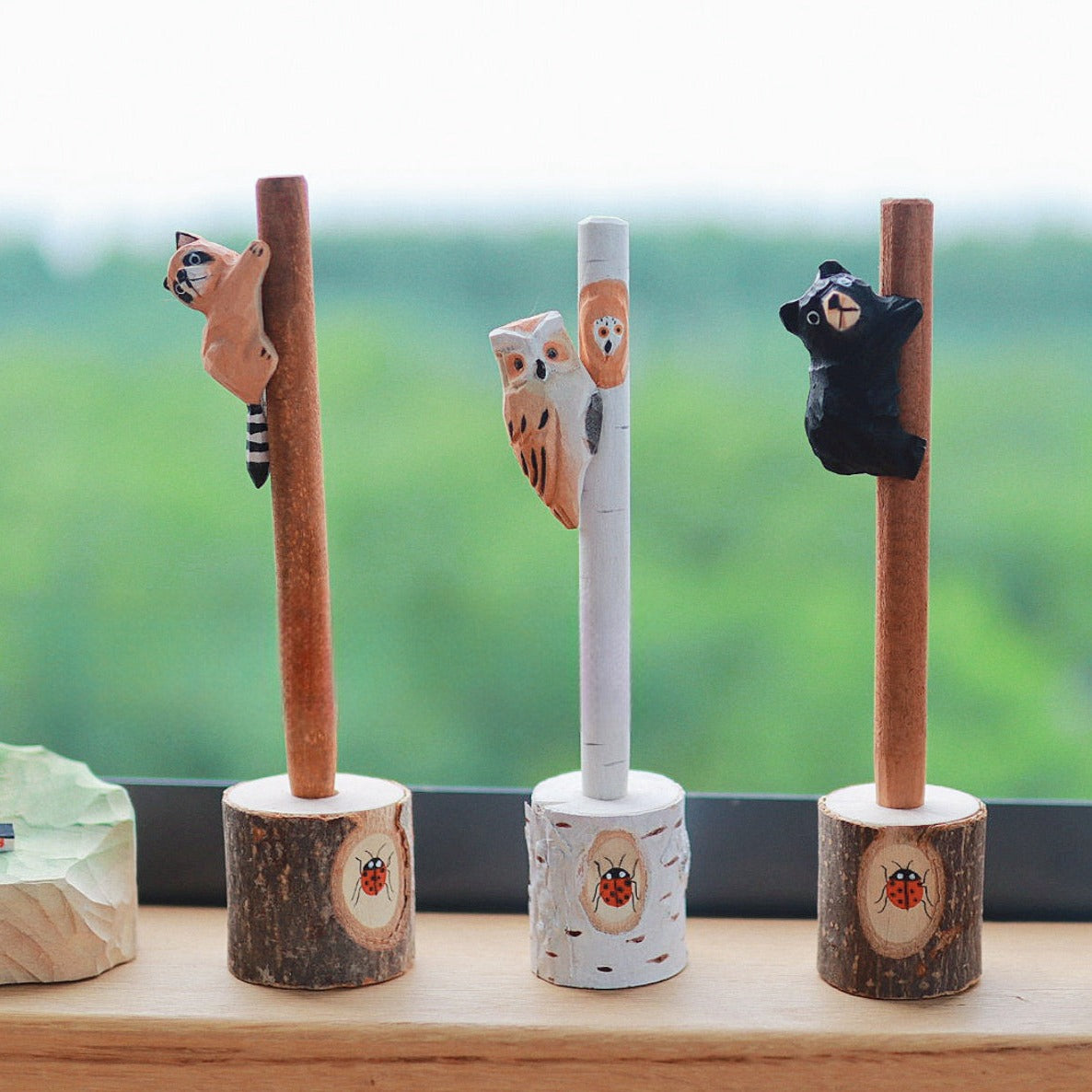 Three wooden pens with hand-carved animal figures (raccoon, owl, and bear) standing upright in tree stump holders.