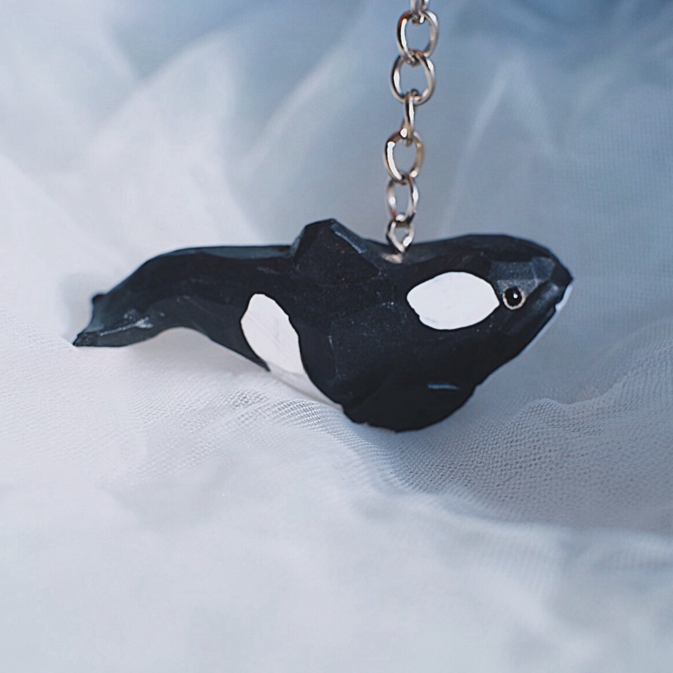 Hand-carved orca whale keychain hanging on a chain, placed on a soft, white fabric background.