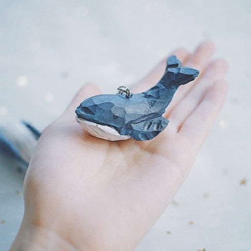 Hand holding a swimming whale keychain, highlighting its intricate carving and blue paint details.